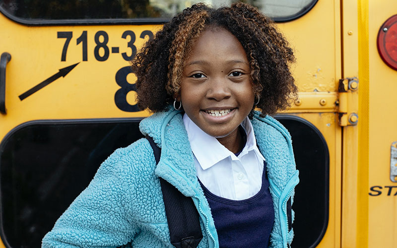 Black child wearing a backpack smiling in front of school bus.