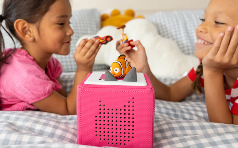 A bright pink digital speaker device known as a Toniebox with a toy figurine on top in the foreground. In the background two little girls sit across from each other listening to the device and playing with two small toy figurines.