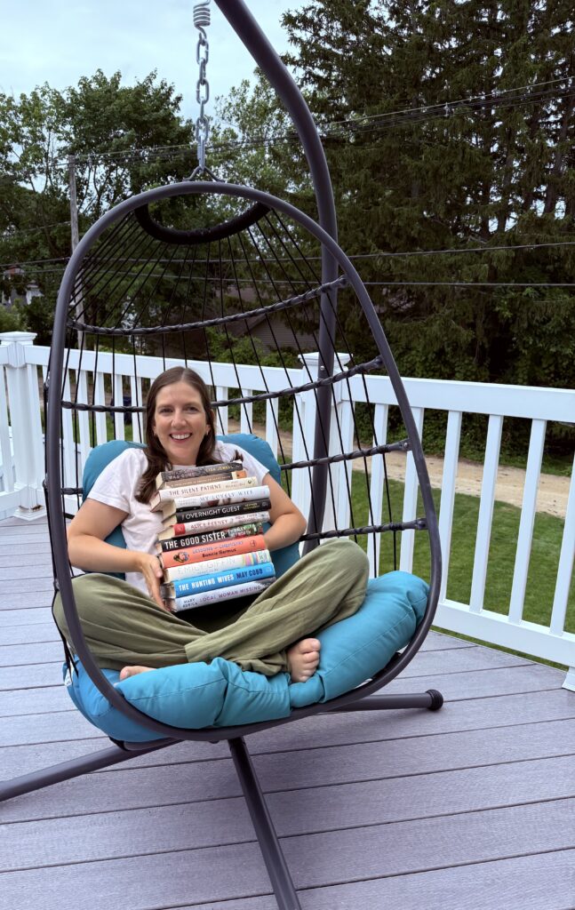 Heather sits on a hanging chair with blue cushions holding a stack of books. 