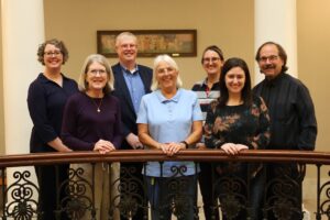 Seven adults pose behind an ornate metal railing in a warmly lit, wood-paneled interior.