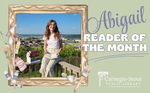 A young girl with brown hair and glasses sits on a railing overlooking Dubuque. She has a stack of books next to her, and an open book on her lap.