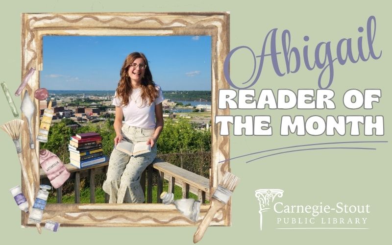 A young girl with brown hair and glasses sits on a railing overlooking Dubuque. She has a stack of books next to her, and an open book on her lap.